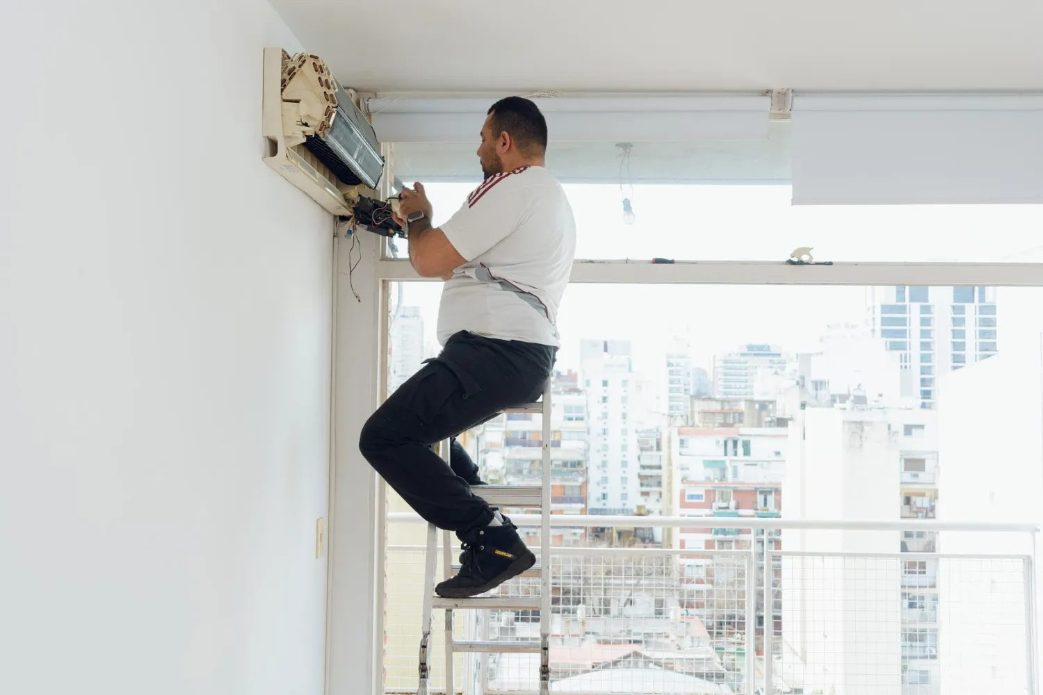 Muž, ktorý na rebríku demontuje klimatizáciu na stene.